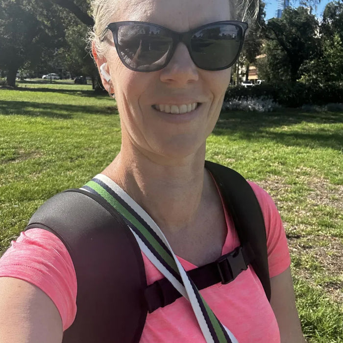 A smiling woman outdoors in a park wearing black sunglasses, a pink shirt, and a black backpack with a green and white strap.