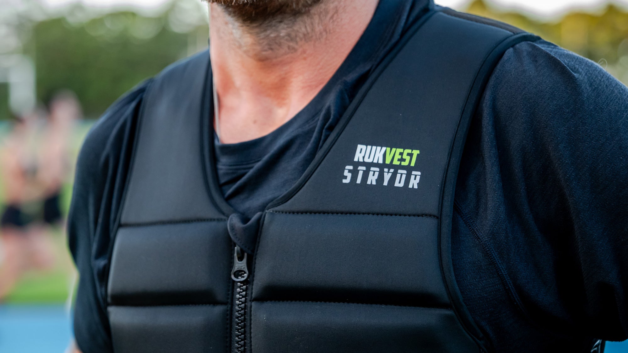 A close-up of a man wearing a black RUKVEST STRYDR running vest over a black t-shirt, with the brand logo printed on the chest.